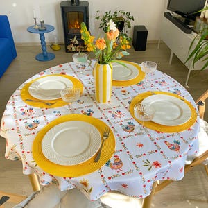 Könnte beinhalten: Ein runder Esstisch für vier Personen, gedeckt mit einer weißen Tischdecke mit bunten Blumen- und Hahnmustern. Jedes Gedeck besteht aus einem weißen Teller, einem gelben Platzset und einem Glas. Eine gelb-weiß gestreifte Vase enthält Blumen.