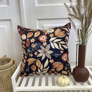 Pode incluir: Uma almofada quadrada com fundo azul-marinho escuro e um padrão repetitivo de flores e folhas em tons de laranja, creme e azul suave. A almofada é exibida ao lado de um vaso castanho e uma pequena abóbora branca.
