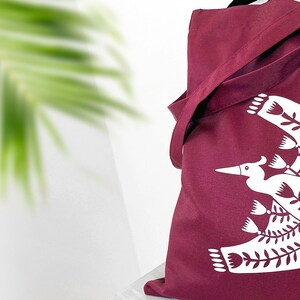 May include: A maroon tote bag with a white design of a stylized bird with floral elements. The design is centered on the bag.