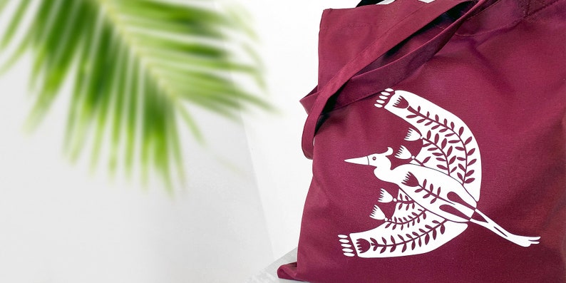 May include: A maroon tote bag with a white design of a stylized bird with floral elements. The design is centered on the bag.