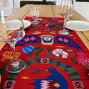 May include: Red table runner with a colorful folk art design featuring birds, flowers, and geometric patterns. The runner is on a wooden table with two wine glasses and plates.