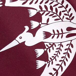 May include: A white stylized bird design on a maroon fabric background. The bird is flying with its wings spread and has a long beak. The design is surrounded by white stylized leaves and flowers.