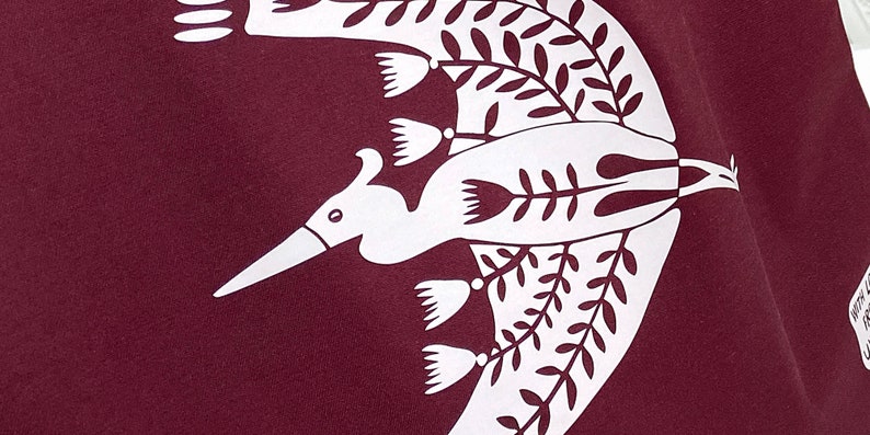 May include: A white stylized bird design on a maroon fabric background. The bird is flying with its wings spread and has a long beak. The design is surrounded by white stylized leaves and flowers.