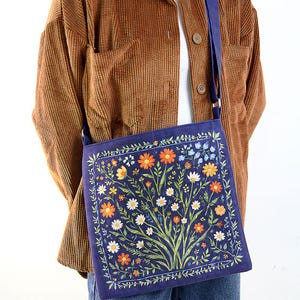 May include: A blue canvas tote bag with a floral print featuring a variety of colorful flowers and leaves. The bag has a long strap and is worn over the shoulder.