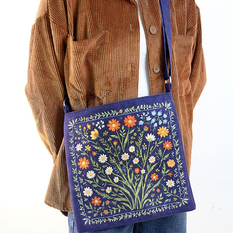 May include: A blue canvas tote bag with a floral print featuring a variety of colorful flowers and leaves. The bag has a long strap and is worn over the shoulder.