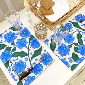Puede incluir: Una mesa decorada con dos manteles individuales florales con flores de hortensia azules y hojas verdes. El conjunto incluye vasos de agua transparentes, una bandeja de mimbre con un plato blanco y posavasos, y un candelabro.