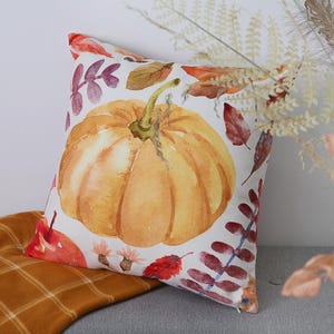 May include: A square decorative pillow with a watercolor pumpkin design. The pumpkin is orange with a green stem, surrounded by fall leaves in shades of red, orange, and purple. The pillow is on a gray surface with a plaid blanket.