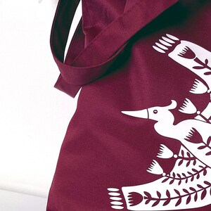 May include: A burgundy tote bag with a white design of a stylized bird with floral elements.