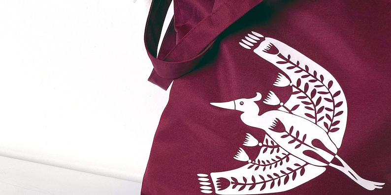 May include: A burgundy tote bag with a white design of a stylized bird with floral elements.