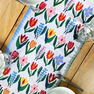 May include: White table runner with a repeating floral pattern in red, orange, pink, yellow, and blue. The flowers have green leaves and are set against a white background. The runner is on a wooden table with plates, forks, and glasses.