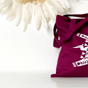 May include: A burgundy tote bag with a white design of a bird flying with branches. The bag has two long straps.