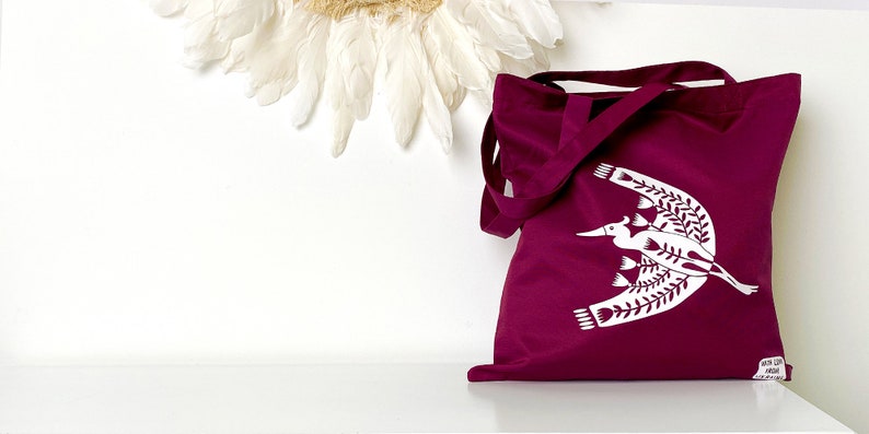 May include: A burgundy tote bag with a white design of a bird flying with branches. The bag has two long straps.