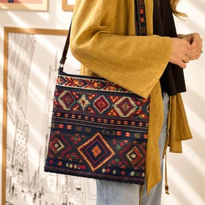 May include: A square crossbody bag featuring a dark blue base with a multi-colored geometric pattern in red, orange, and green. The bag has a long, thin dark strap and a repeating diamond and floral motif across the front surface of the fabric.