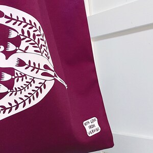 May include: A purple tote bag with a white bird design and floral accents. The bag has a white label that says "With Love From Ukraine".