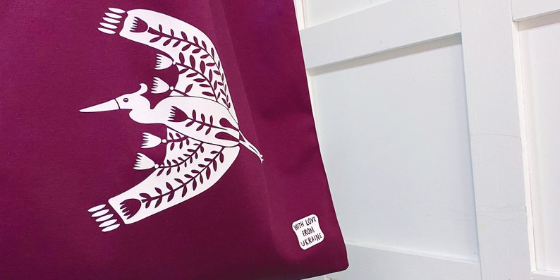 May include: A purple tote bag with a white bird design and floral accents. The bag has a white label that says "With Love From Ukraine".