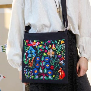 May include: Black crossbody bag with a vibrant, embroidered floral design. The embroidery features colorful flowers, birds, and a fox. The bag has a long, adjustable strap.