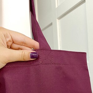 May include: A burgundy tote bag with a single strap held by a hand. The bag has a stitched seam across the top.