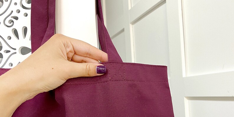 May include: A burgundy tote bag with a single strap held by a hand. The bag has a stitched seam across the top.