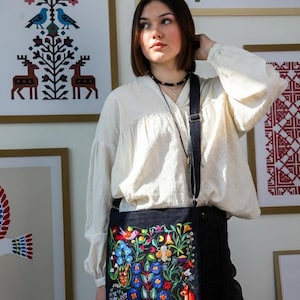 May include: A black crossbody bag with a vibrant floral embroidery design featuring blue, red, and yellow flowers, birds, and a fox. The bag has a long, adjustable strap and is worn over the shoulder. The background includes framed artwork.