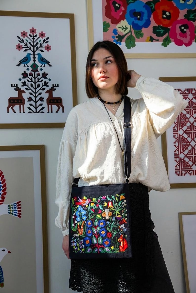May include: A black crossbody bag with a vibrant floral embroidery design featuring blue, red, and yellow flowers, birds, and a fox. The bag has a long, adjustable strap and is worn over the shoulder. The background includes framed artwork.