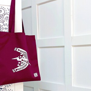 May include: A burgundy tote bag with a white bird design. The bag has a white handle and a white tag on the bottom right corner.