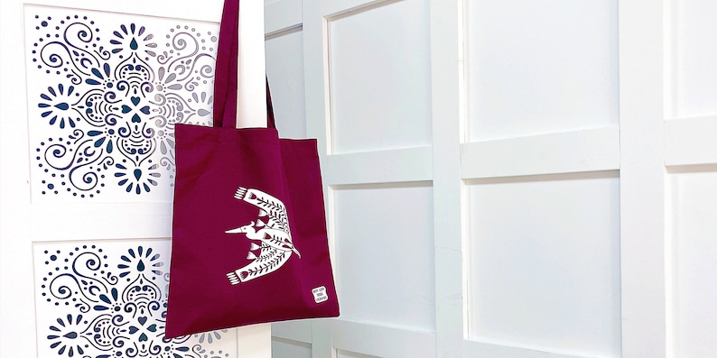 May include: A burgundy tote bag with a white bird design. The bag has a white handle and a white tag on the bottom right corner.