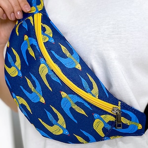 May include: A blue and yellow fanny pack with a zipper closure and a pattern of stylized birds. The fanny pack is worn around the waist.
