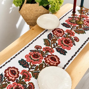 Puede incluir: Un camino de mesa blanco con un patrón floral repetitivo en tonos rojo, rosa y verde oscuro. El camino está colocado sobre una mesa de madera clara con dos cuencos de cerámica color crema, una cesta tejida con follaje y dos velas altas.