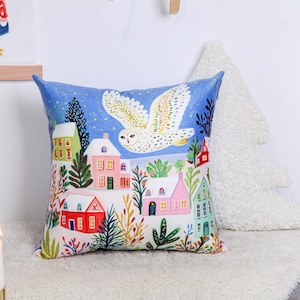 Może przedstawiać: Dekoracyjna kwadratowa poduszka z fantazyjną zimową scenerią. Poduszka ma niebieskie tło z latającą sową śnieżną nad kolorowymi domami. Domy mają czerwone, różowe, zielone i żółte dachy. Poduszka leży na białej powierzchni.