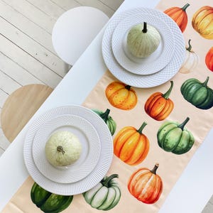 Può includere: Un runner da tavola rettangolare con una stampa ad acquerello di zucche nei toni dell'arancione, giallo e verde su sfondo beige chiaro. Due piatti in ceramica bianca sono posizionati sul runner, ciascuno sormontato da una piccola zucca verde pallido.