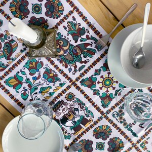 May include: A white table runner with a vibrant folk art pattern of birds, flowers, and figures. The runner is set with a candle holder, plates, cutlery, and glassware, creating a decorative table setting.