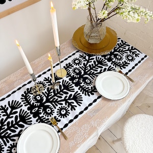 May include: A dining table set for a meal, featuring a white and black floral patterned table runner. Two white plates with black rims are set with gold forks. Tall white candles in silver holders and a vase of white flowers complete the setting.