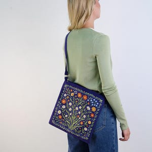May include: A purple shoulder bag with a floral embroidery design. The bag has a white strap and is worn by a person with blonde hair.