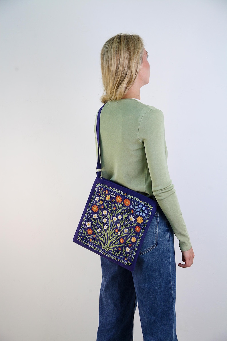 May include: A purple shoulder bag with a floral embroidery design. The bag has a white strap and is worn by a person with blonde hair.