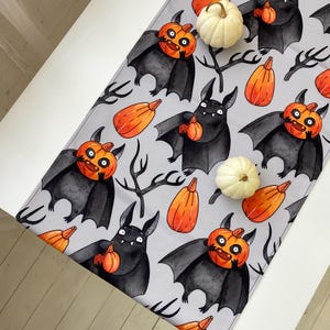 Könnte beinhalten: Ein hellgrauer Tischläufer mit einem sich wiederholenden Muster aus schwarzen Fledermäusen, die orangefarbene Kürbisse halten, durchsetzt mit orangefarbenen Kürbissen und schwarzen, blattlosen Zweigen. Zwei kleine, weiße Zierkürbisse liegen auf dem Stoff.