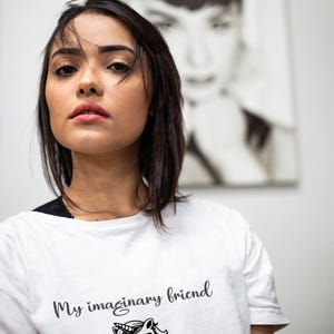 May include: White t-shirt with the text "My imaginary friend that I need a". The shirt features a black and white illustration of an angry unicorn. A black and white portrait is in the background.