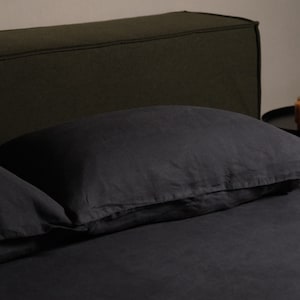 May include: Dark gray linen bedding set on a bed. The set includes a pillow and a fitted sheet. The headboard is a dark green color. A small black side table with a wooden cup is in the background.