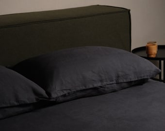 Könnte beinhalten: Dunkelgraues Leinenbettwäsche-Set auf einem Bett. Das Set beinhaltet ein Kissen und ein Spannbettlaken. Das Kopfteil ist dunkelgrün. Ein kleiner schwarzer Beistelltisch mit einem Holzbecher befindet sich im Hintergrund.