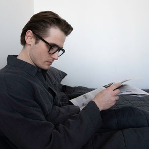 May include: A person wearing black-rimmed glasses sits in bed reading a magazine. They are wearing a dark grey button-down shirt and are covered by a dark grey quilted blanket.