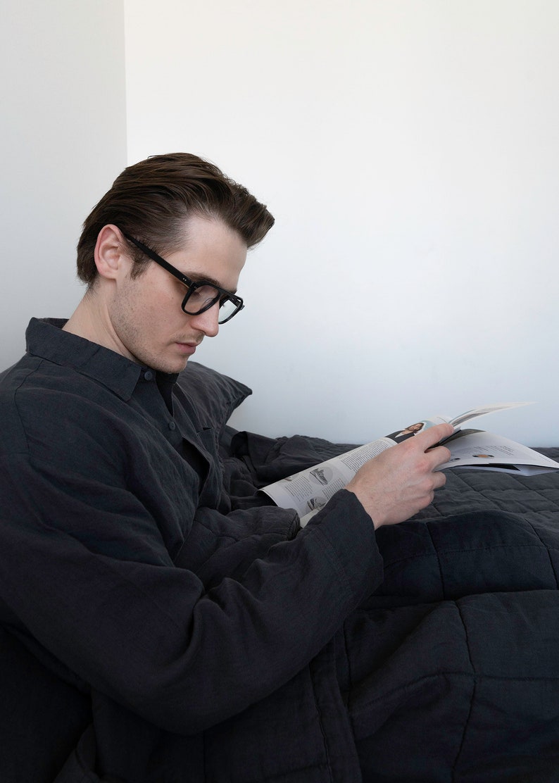 May include: A person wearing black-rimmed glasses sits in bed reading a magazine. They are wearing a dark grey button-down shirt and are covered by a dark grey quilted blanket.