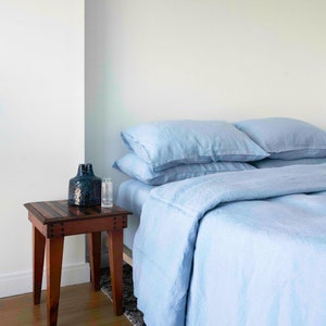 Puede incluir: Un juego de cama de lino azul claro con dos almohadas en una cama. Una mesita de noche de madera con un jarr&oacute;n azul oscuro y una botella de vidrio est&aacute; al lado de la cama.