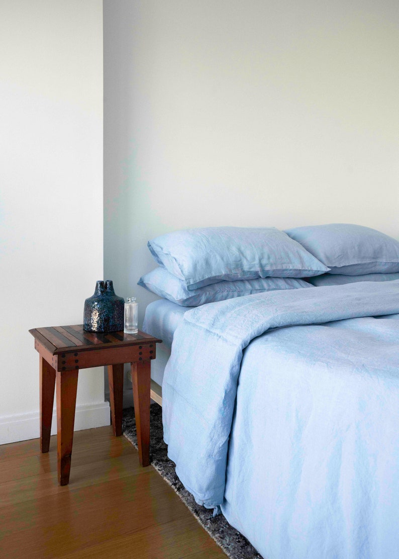 Puede incluir: Un juego de cama de lino azul claro con dos almohadas en una cama. Una mesita de noche de madera con un jarr&oacute;n azul oscuro y una botella de vidrio est&aacute; al lado de la cama.