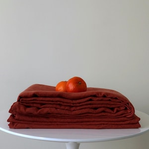 May include: A stack of rust-colored linen fabric folded neatly on a white pedestal table. Two small orange clementines rest on top of the fabric.