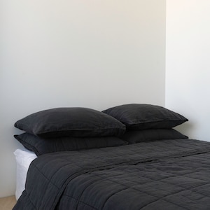 May include: A black linen quilt with a quilted pattern. The quilt is folded on a bed with two black linen pillows.
