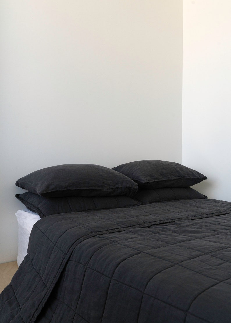 May include: A black linen quilt with a quilted pattern. The quilt is folded on a bed with two black linen pillows.