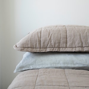 May include: A close-up of a beige quilted bedspread with a light grey pillow on top. The bedspread has a square quilted pattern.