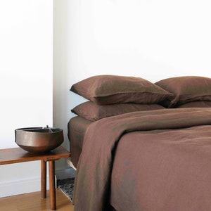 Puede incluir: Una funda n&oacute;rdica y fundas de almohada de lino marr&oacute;n sobre una cama con una mesita de noche de madera marr&oacute;n y un cuenco de metal en la parte superior.