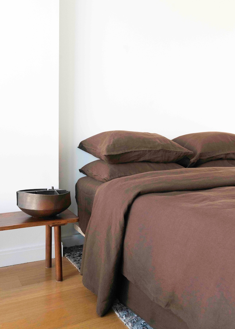 Puede incluir: Una funda n&oacute;rdica y fundas de almohada de lino marr&oacute;n sobre una cama con una mesita de noche de madera marr&oacute;n y un cuenco de metal en la parte superior.