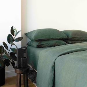 Puede incluir: Una funda n&oacute;rdica y fundas de almohada de lino verde en una cama con un taburete de madera y una planta de interior en la esquina.