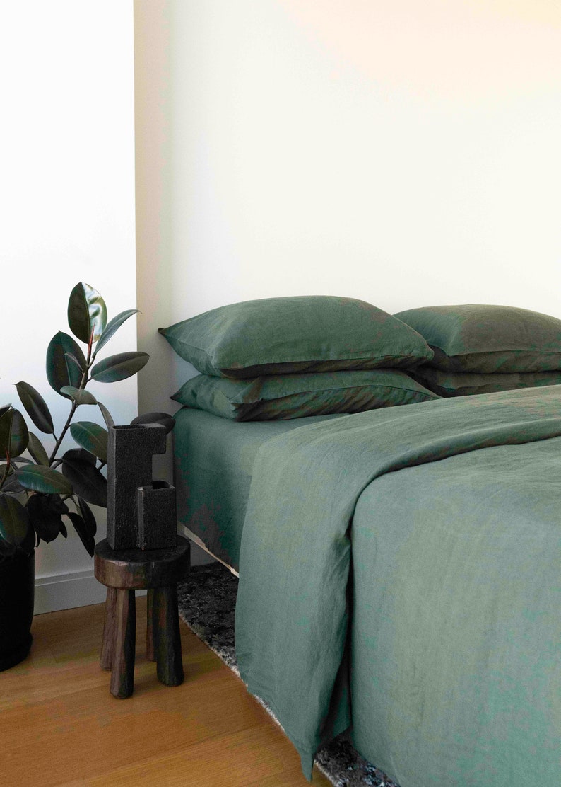 Puede incluir: Una funda n&oacute;rdica y fundas de almohada de lino verde en una cama con un taburete de madera y una planta de interior en la esquina.
